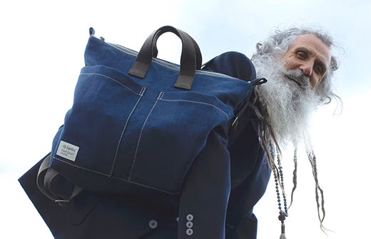 Stewart Gilchrist, a man with grey hair and plaits in his beard, looks down at the camera with the sky behind him. He wears a blue backpack with grey handles